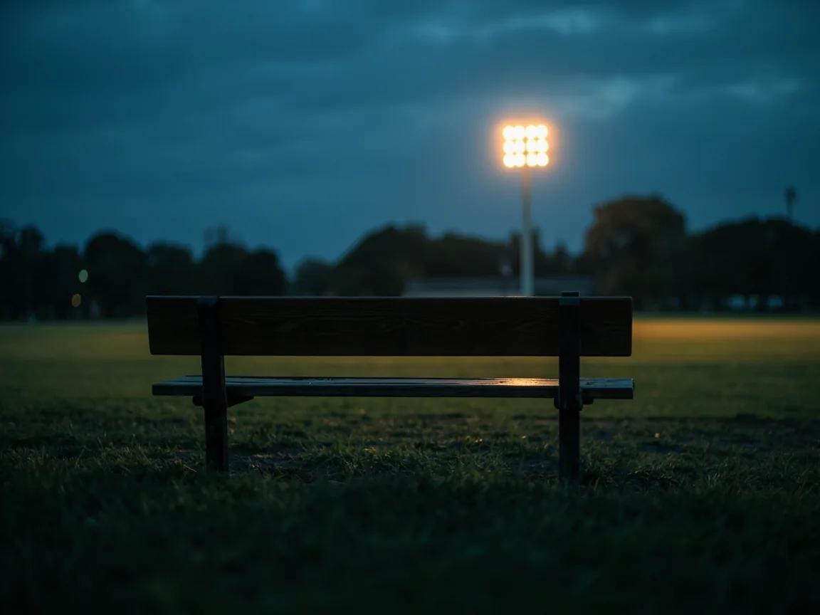 Editorial still of a single wooden park bench beside an empty cricket oval at dusk, soft floodlight glow on the horizon — editorial scope card.